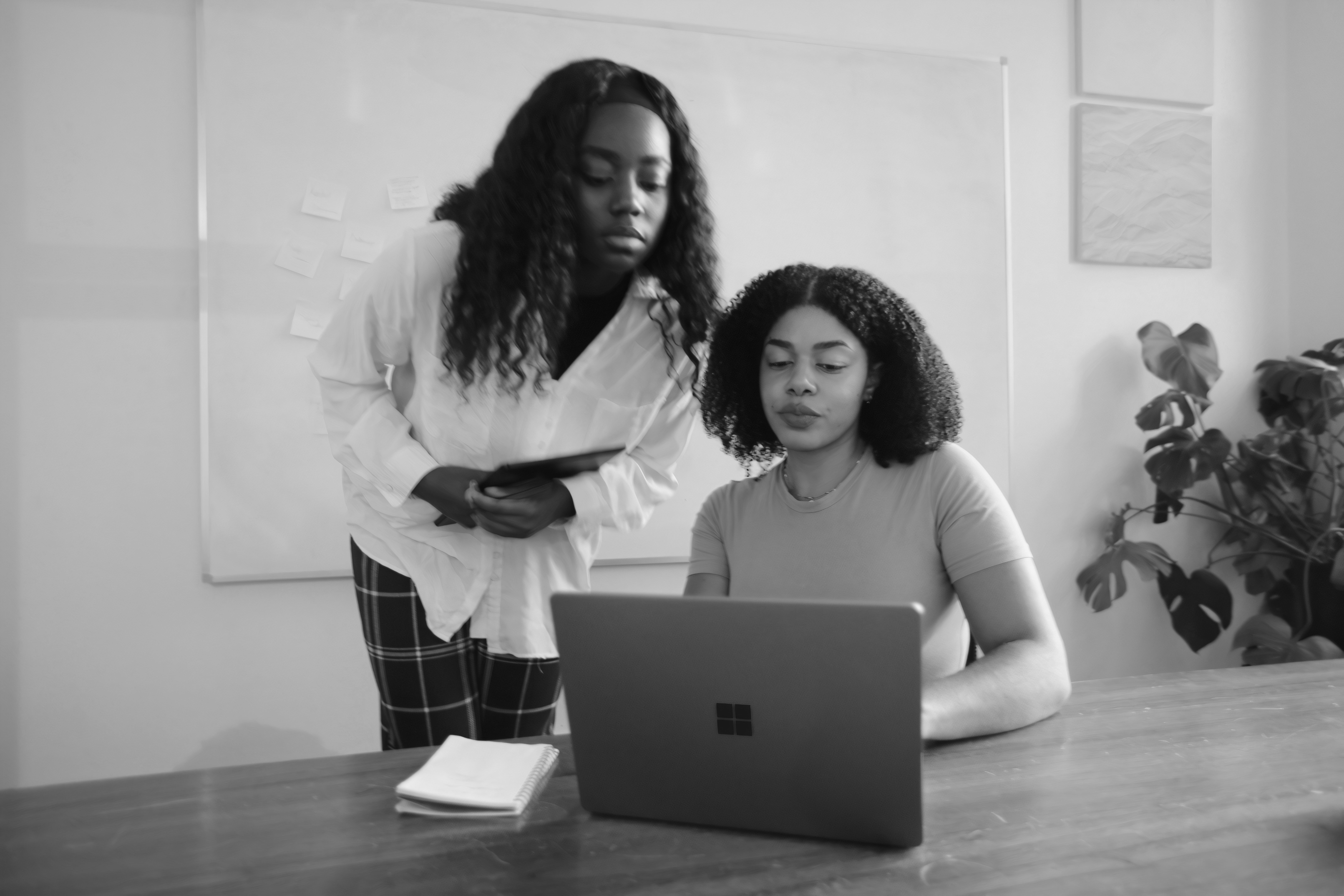Two women in a workspace: one standing, observing, while the other sits at a laptop, focused on her work.