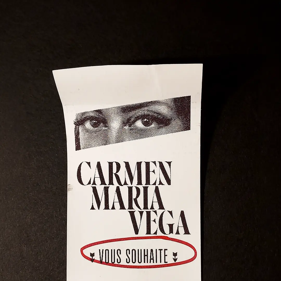 Carte de vœux verticale pour l'artiste Carmen Maria Vega. Le visuel présente un gros plan en noir et blanc sur un regard intense, surmontant le nom de l'artiste en typographie serif élégante. En bas, l'inscription 'VOUS SOUHAITE' est entourée d'un trait rouge.