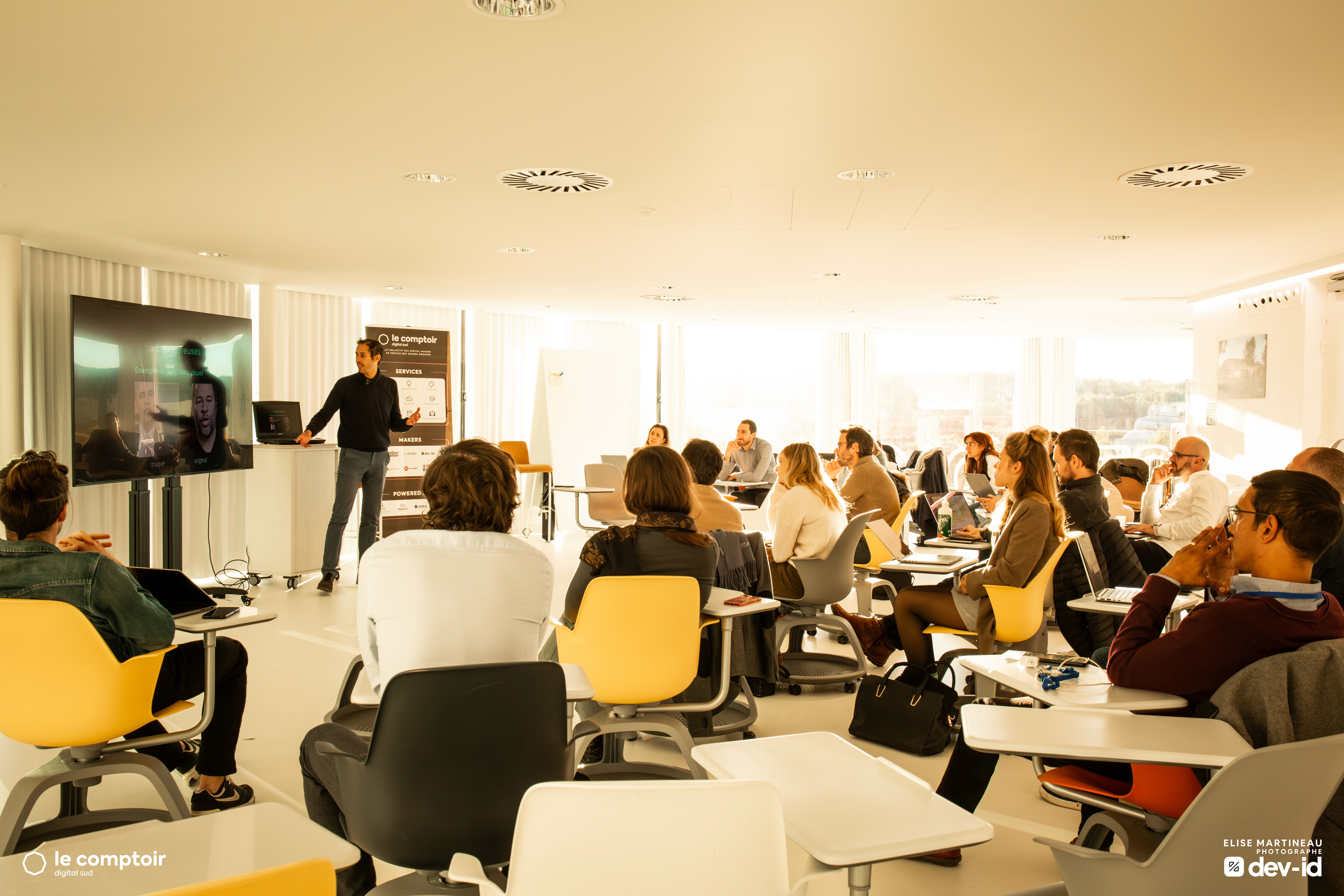 Un groupe de personnes assises dans une salle lumineuse, participant à une présentation dirigée par un homme qui se tient debout près d’un écran.