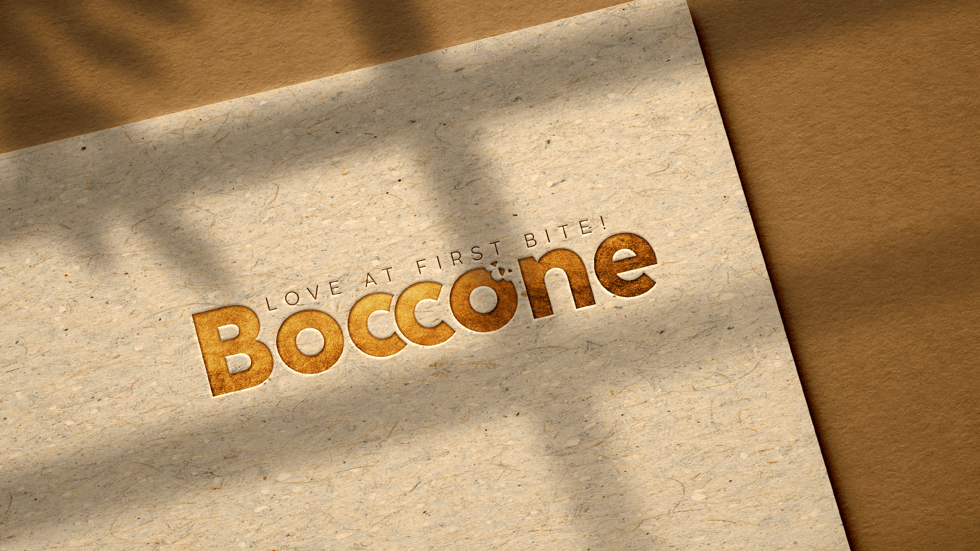 Embossed Boccone Café logo with the tagline ‘Love at First Bite’ on textured kraft paper, lit with soft natural shadows.