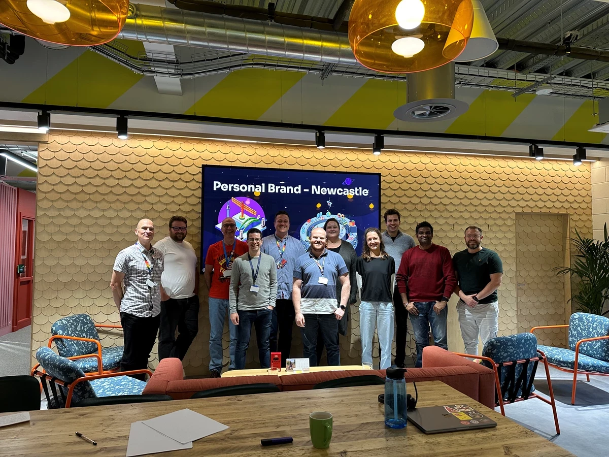 Group of people standing in front of a screen that reads “Personal Brand – Newcastle” in a modern meeting space with bright lighting and colorful chairs.