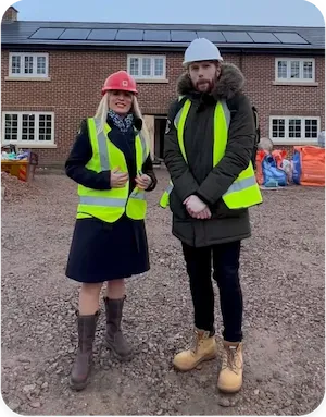 Two people wearing high-visibility vests and hard hats standing on a construction site in front of a new brick house development