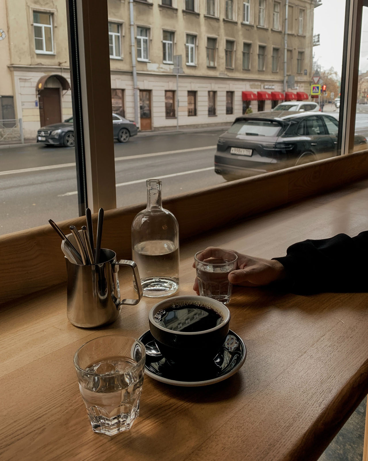 A hand holding a glass of water next to a black coffee looking onto the street