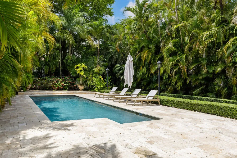 Rectangular stone-tiled backyard pool surrounded by dense tropical greenery and sun loungers.