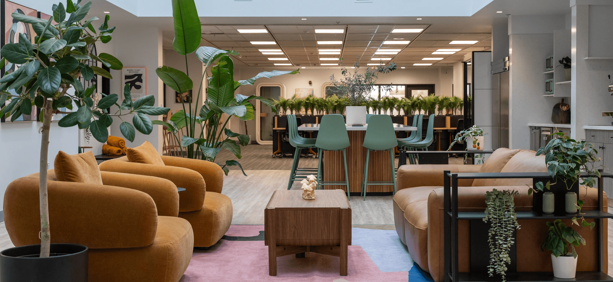 Bright open-plan co-working space filled with lush plants, wooden desks, and a person working at a computer by floor-to-ceiling windows