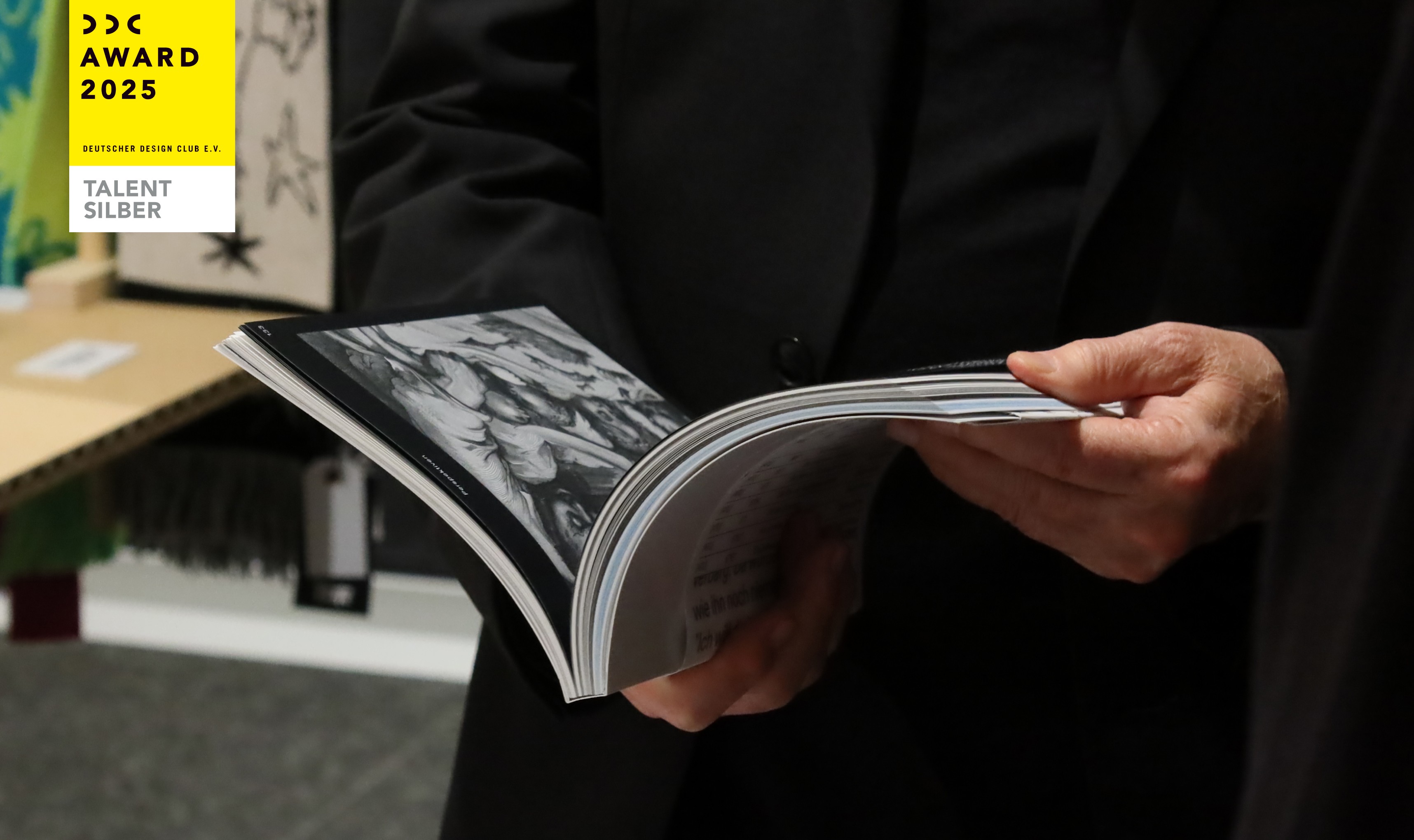 A person holds an open book featuring black and white illustrations, exemplifying editorial design and visual identity in a purpose-driven branding context, against the backdrop of an award display in Germany.