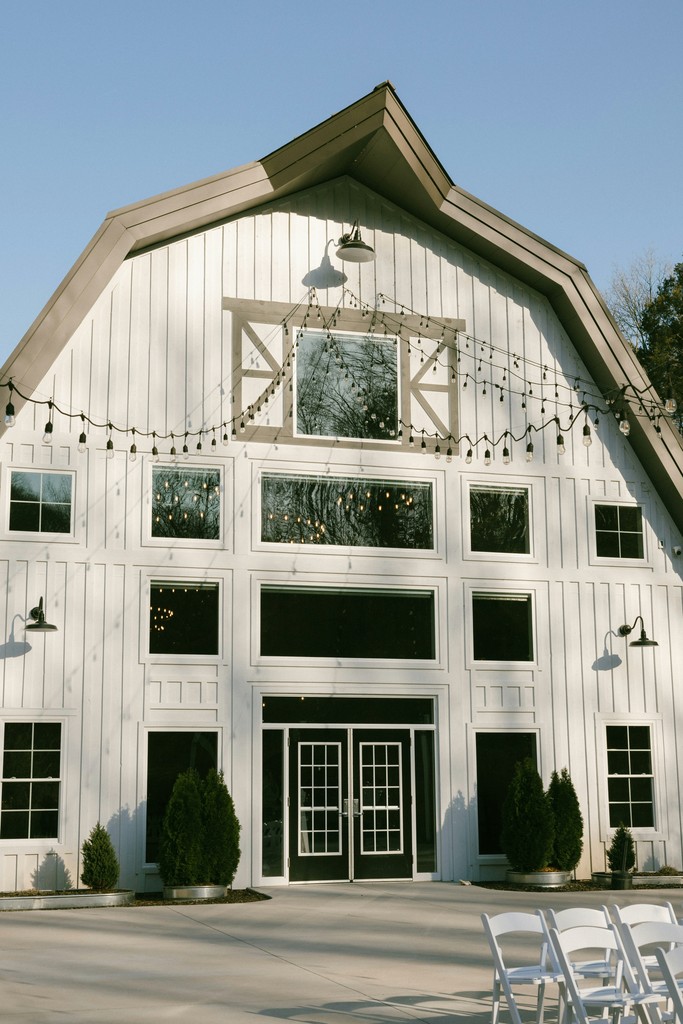 White gambrel barn wedding venue facade with large windows, string lights, farmhouse sconces, and evergreen topiaries by the entrance
