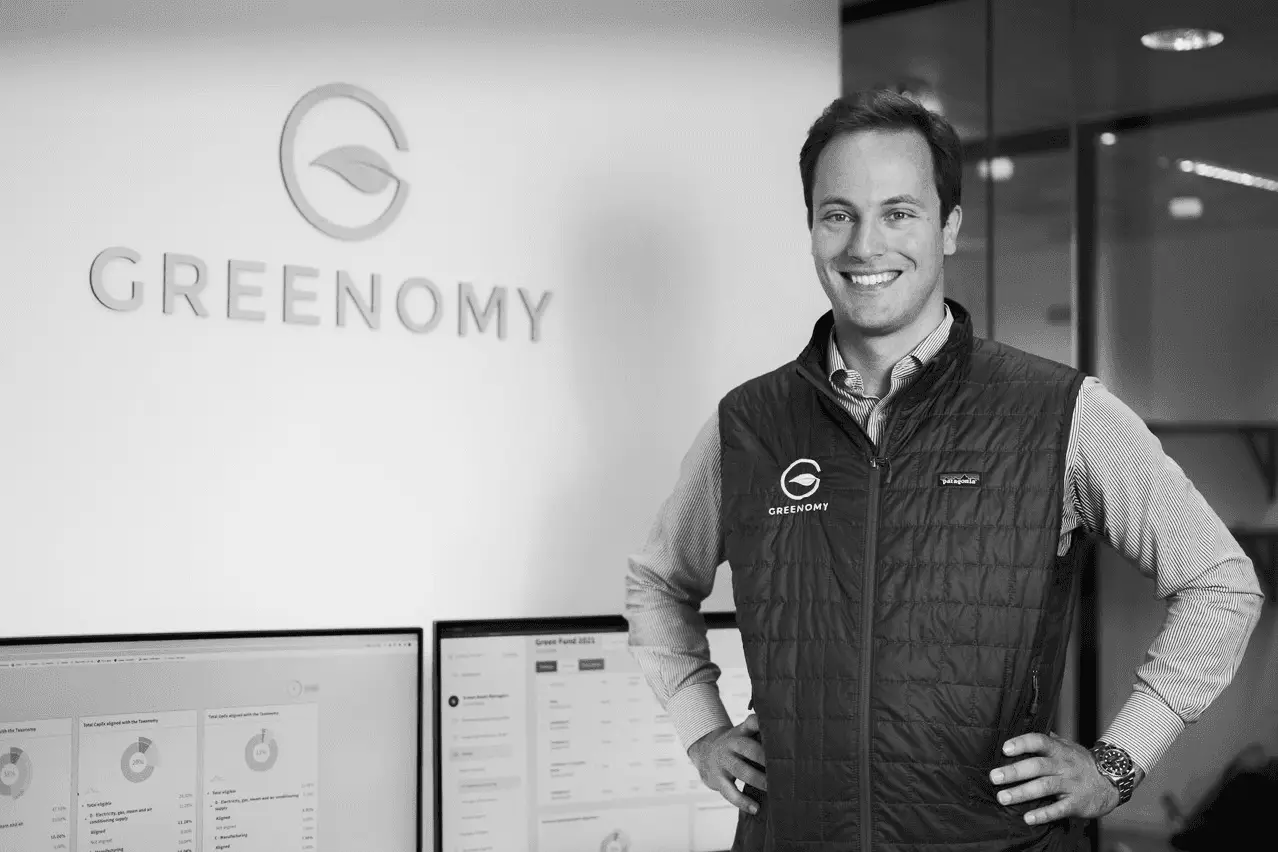 A black and white photo of a smiling professional man standing in an office in front of a "GREENONOMY" logo.