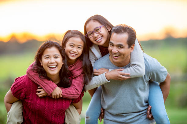 A smiling family of four poses together outdoors, with two children on the parents' backs, enjoying a sunset.