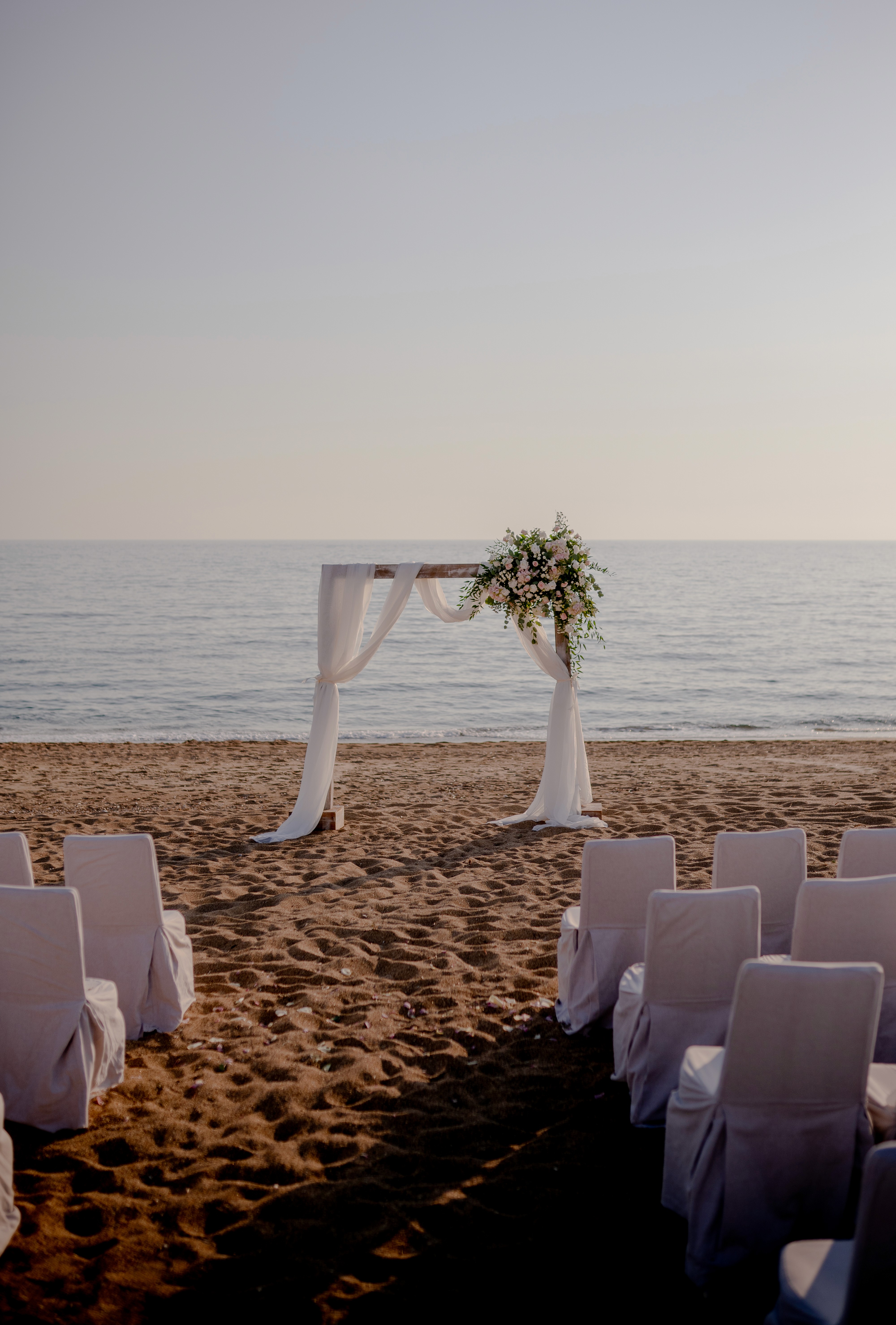 Ιδιαίτερος στολισμός τελετής γάμου στην παραλία με λευκές καρέκλες και αψίδα λουλουδιών