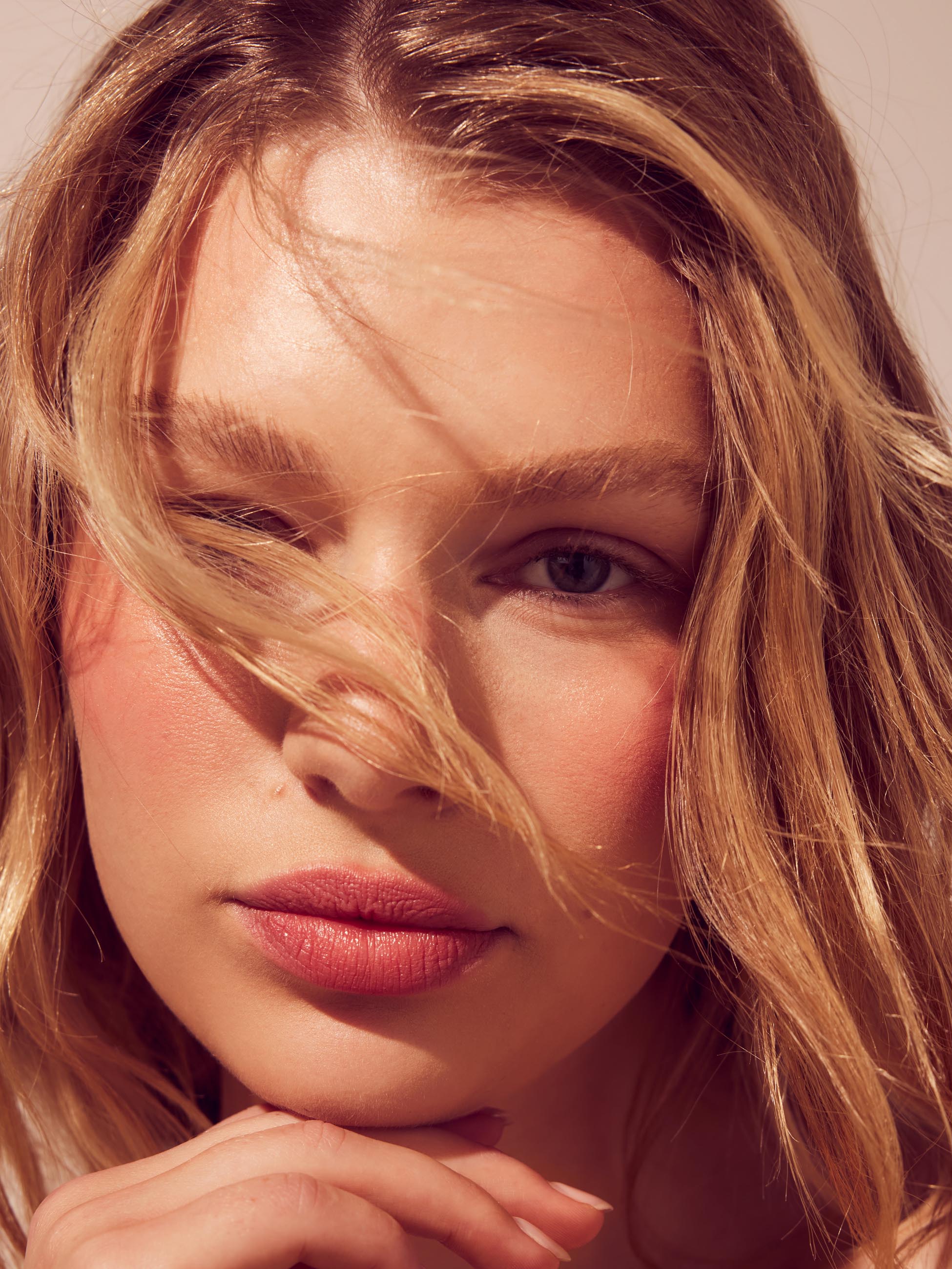 A close-up photo of a blonde woman's face in strong, warm sunshine. Her chin is resting on the back of her hand and strands of her hair are blowing across her face.