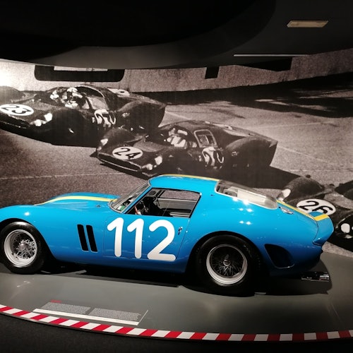 A vintage blue race car with the number 112 displayed in front of a black-and-white photo of race cars on a track.