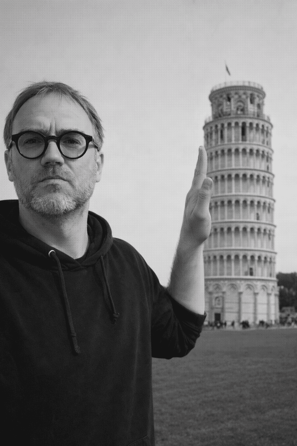 Man with short hair and beard wearing a dark blazer and black shirt against a plain background.