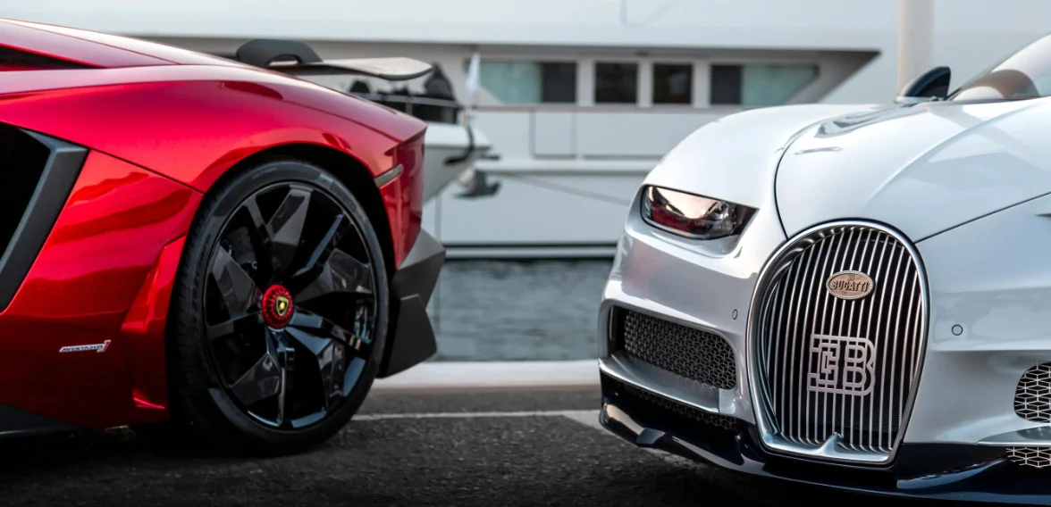 Un face-à-face entre deux supercars d’exception : l’aile arrière et la roue noire d’une sportive rouge contrastent avec la face avant sculptée d’une Bugatti blanche. L’image capture un instant rare où deux visions extrêmes de la performance se croisent dans un décor portuaire haut de gamme.