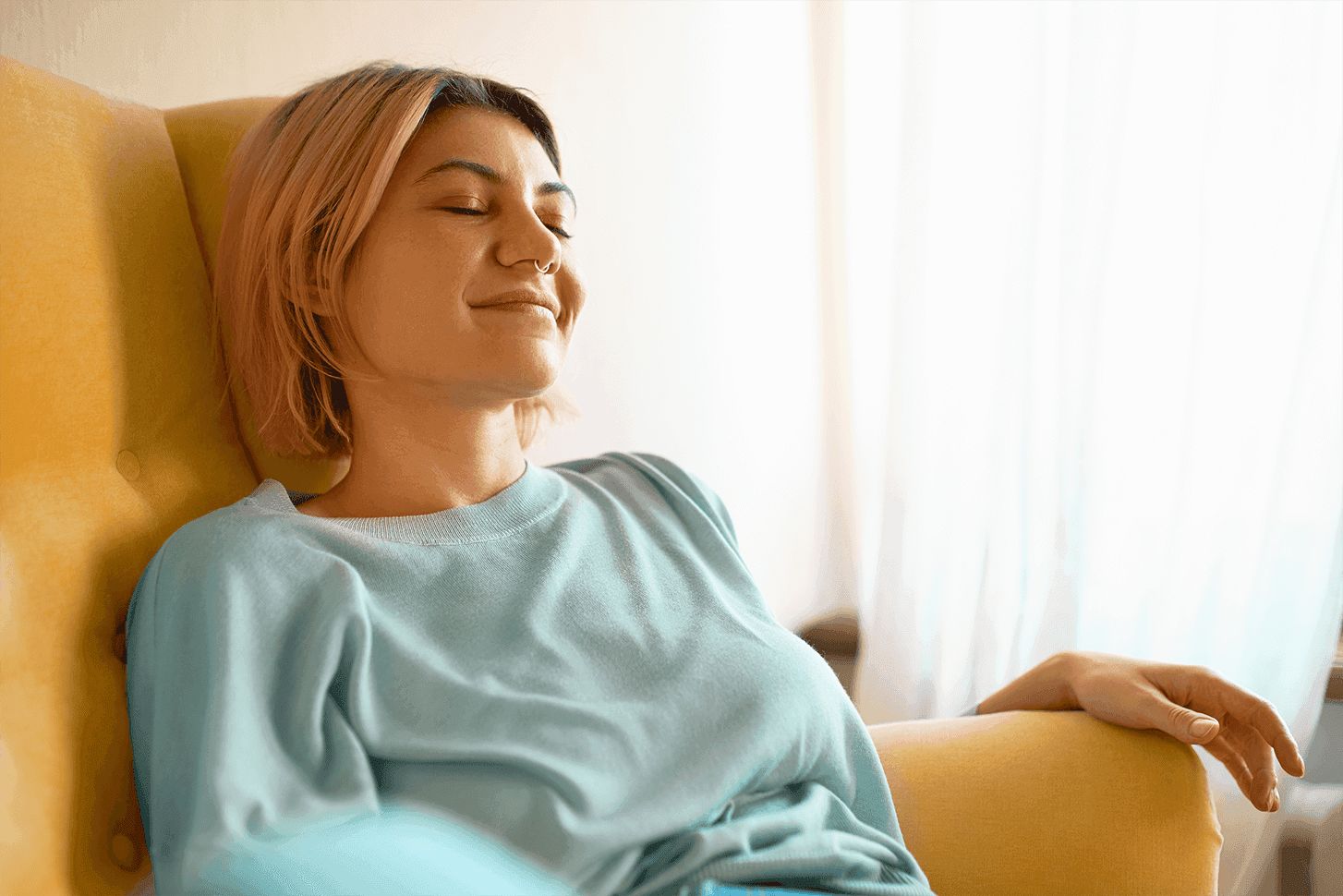 Woman relaxing with eyes closed while sitting comfortably in an armchair in a bright room.