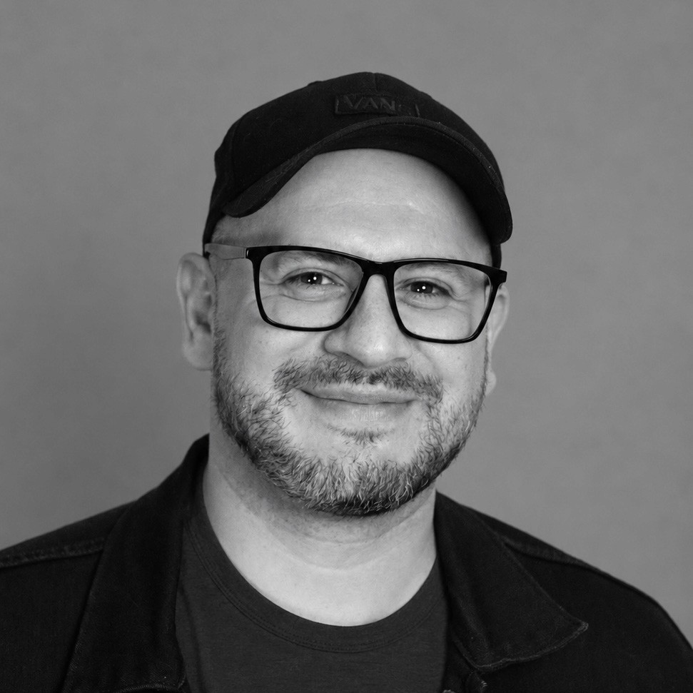 Artistic portrait of a man wearing glasses, illuminated by a diagonal streak of light against a gray background.