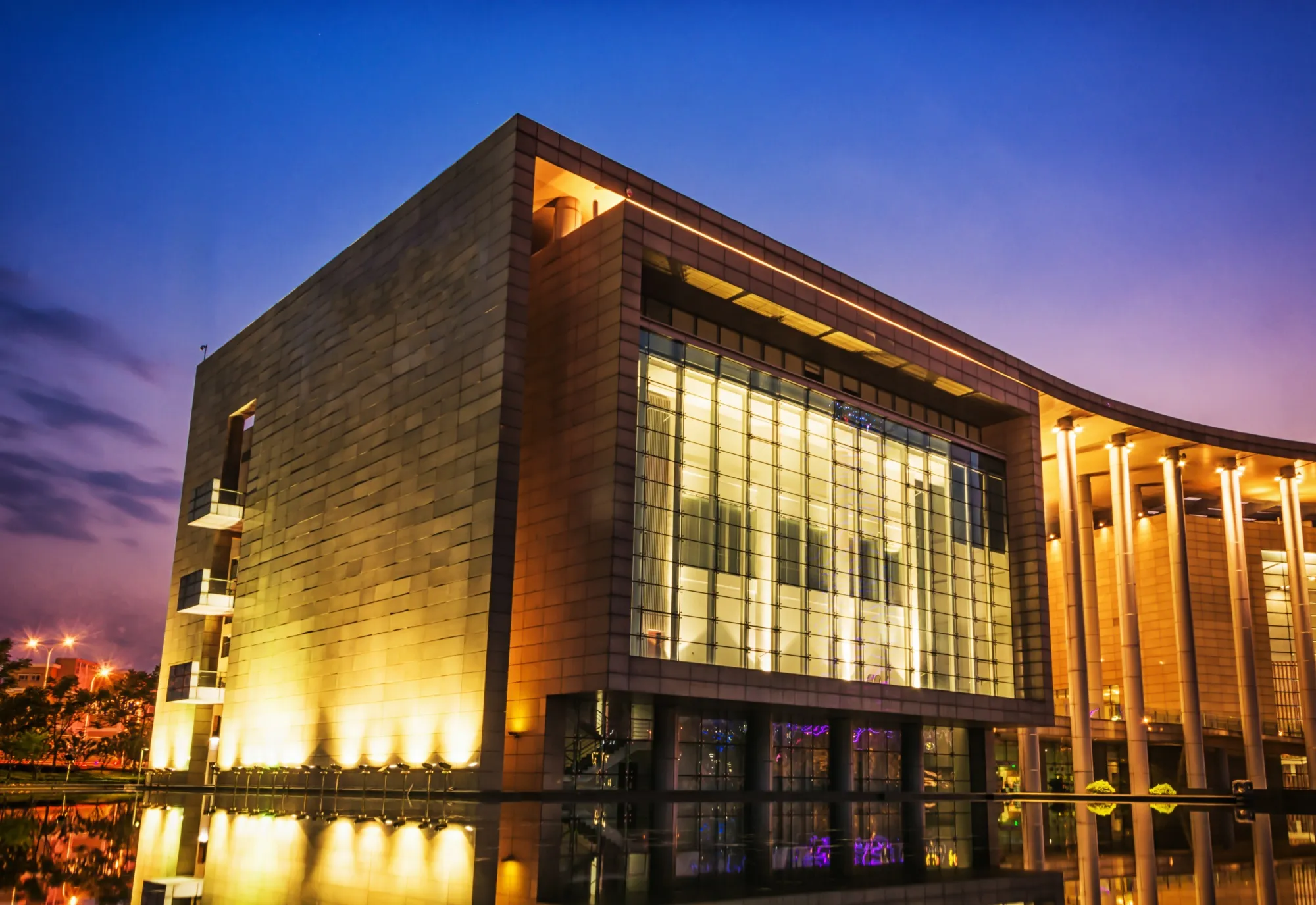 Modern commercial building with illuminated glass facade reflecting on a quiet water feature at twilight.
