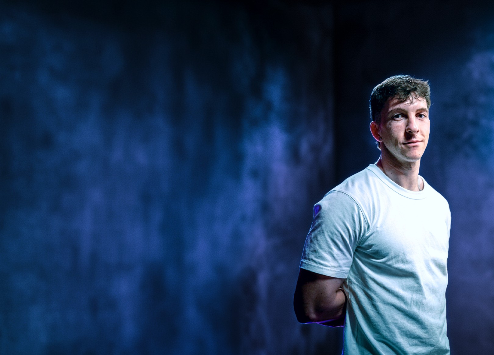 Portrait professionnel et moderne d'un jeune homme, représentant le fondateur ou directeur artistique de Nux-Studio, posant de face avec un léger sourire. Il porte un simple t-shirt blanc à manches courtes. L'arrière-plan est un mur texturé, sombre et baigné d'un éclairage dramatique de couleur bleu électrique et violet, créant une ambiance de studio créatif et innovante. L'espace négatif important sur la gauche est idéal pour l'ajout d'un titre, d'un logo, ou d'un slogan. Ce visuel évoque l'expertise en branding, direction artistique et l'identité moderne d'une agence de design digital.