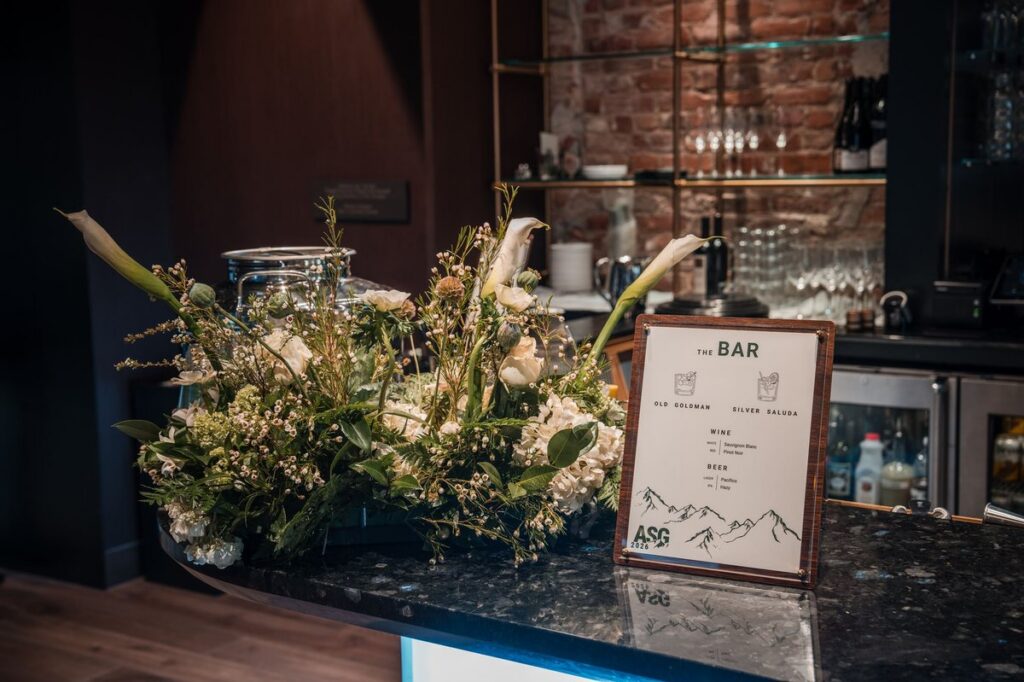 Paul JAS Center bar counter with large floral arrangement, branded cocktail menu, and brass-shelved back bar against exposed brick