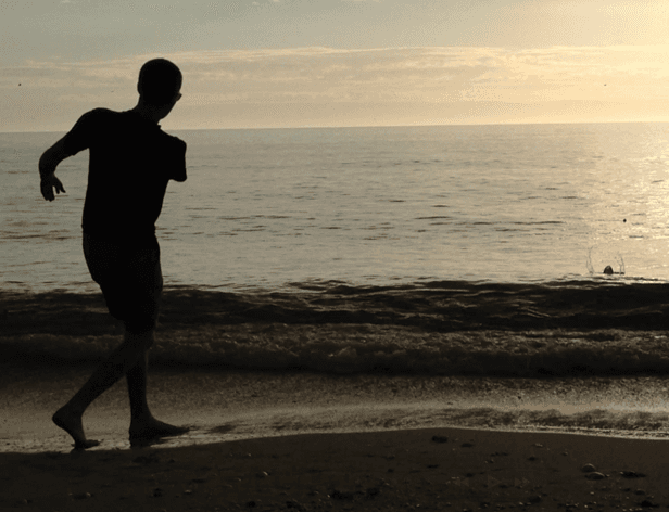 A silhouetted figure is captured mid-stride on a beach, moving away from the viewer towards the ocean. The figure is wearing a short-sleeved shirt and shorts, and their feet are positioned on the wet sand near the water's edge. The ocean stretches out behind them, with gentle waves visible near the shore. The sky above is a light gray or beige color, suggesting an overcast day or soft lighting conditions. The image is in sepia or monochrome tones, giving it a warm tone.