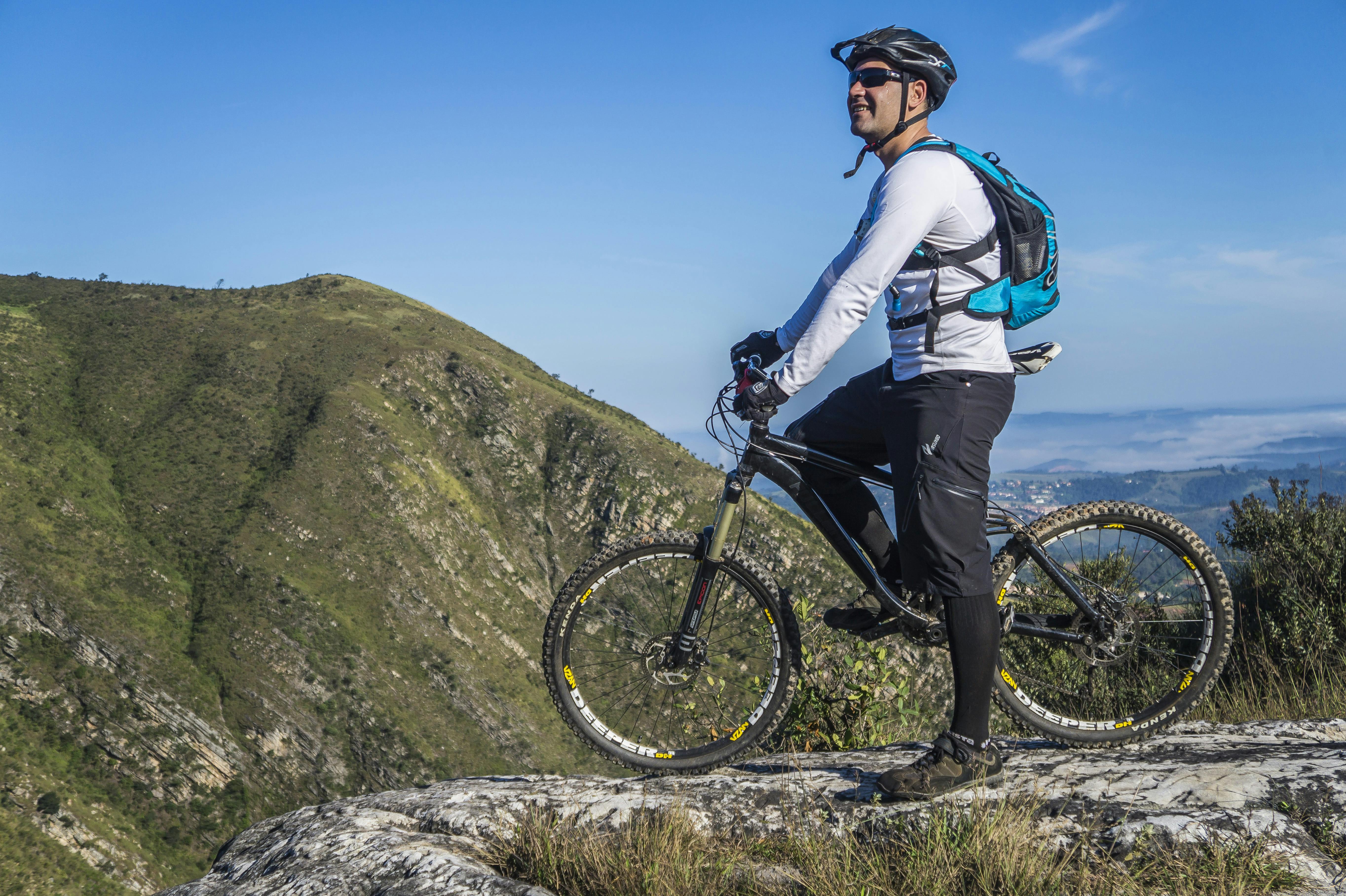 Cyclist standing with a mountain bike on a rocky ridge overlooking green hills.