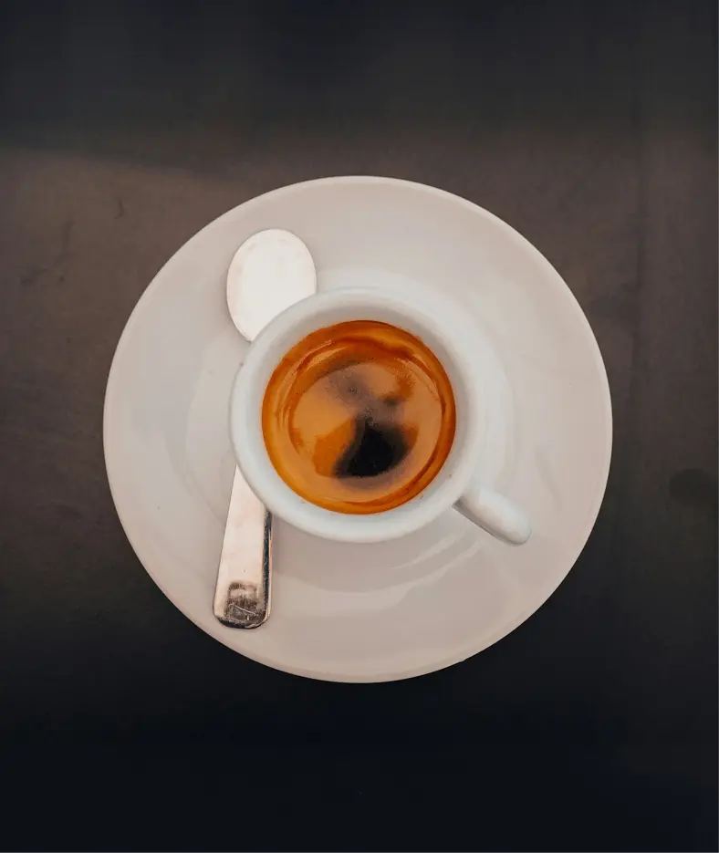 A rich, dark espresso served in a small white cup on a saucer, with a shiny silver spoon resting beside it, set against a dark tabletop background.
