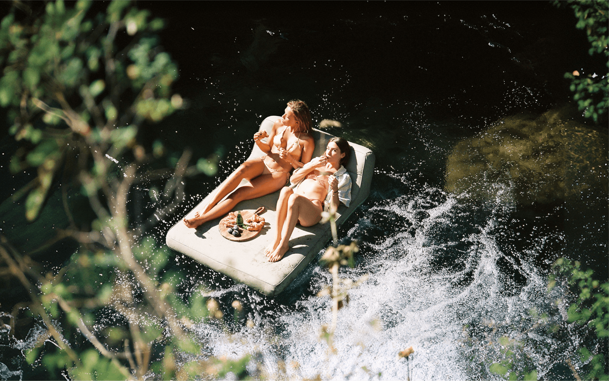 Two women drift lazily on a luxury pool float on the sun-dappled Gorges du Loup river, Côte d'Azur, snacking from a wooden board surrounded by lush greenery.