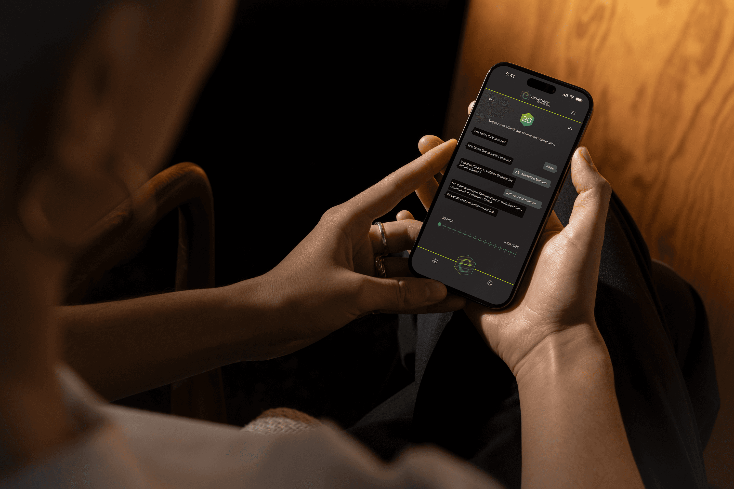 Over-the-shoulder view of a person holding a smartphone using Experteer's interactive conversational onboarding chat interface in a warm, low-light interior setting.