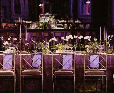 Elegantly set dining table with chairs, adorned with flowers and candles, illuminated by soft purple lighting.