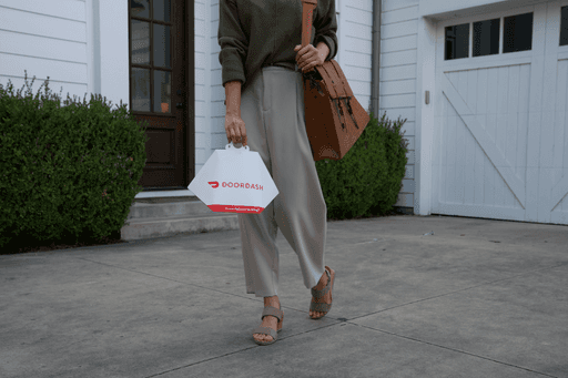 A person carrying a DoorDash takeout bag walks on a concrete driveway, wearing beige pants, brown sandals, and carrying a brown handbag, with a white house and garage in the background.