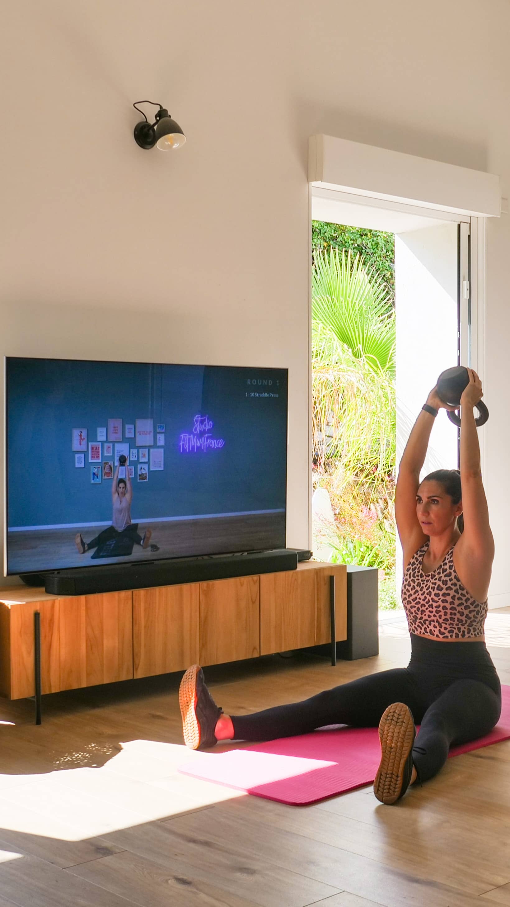 Maman réalisant une séance de sport à la maison avec un kettlebell devant son programme FitMum sur la télévision