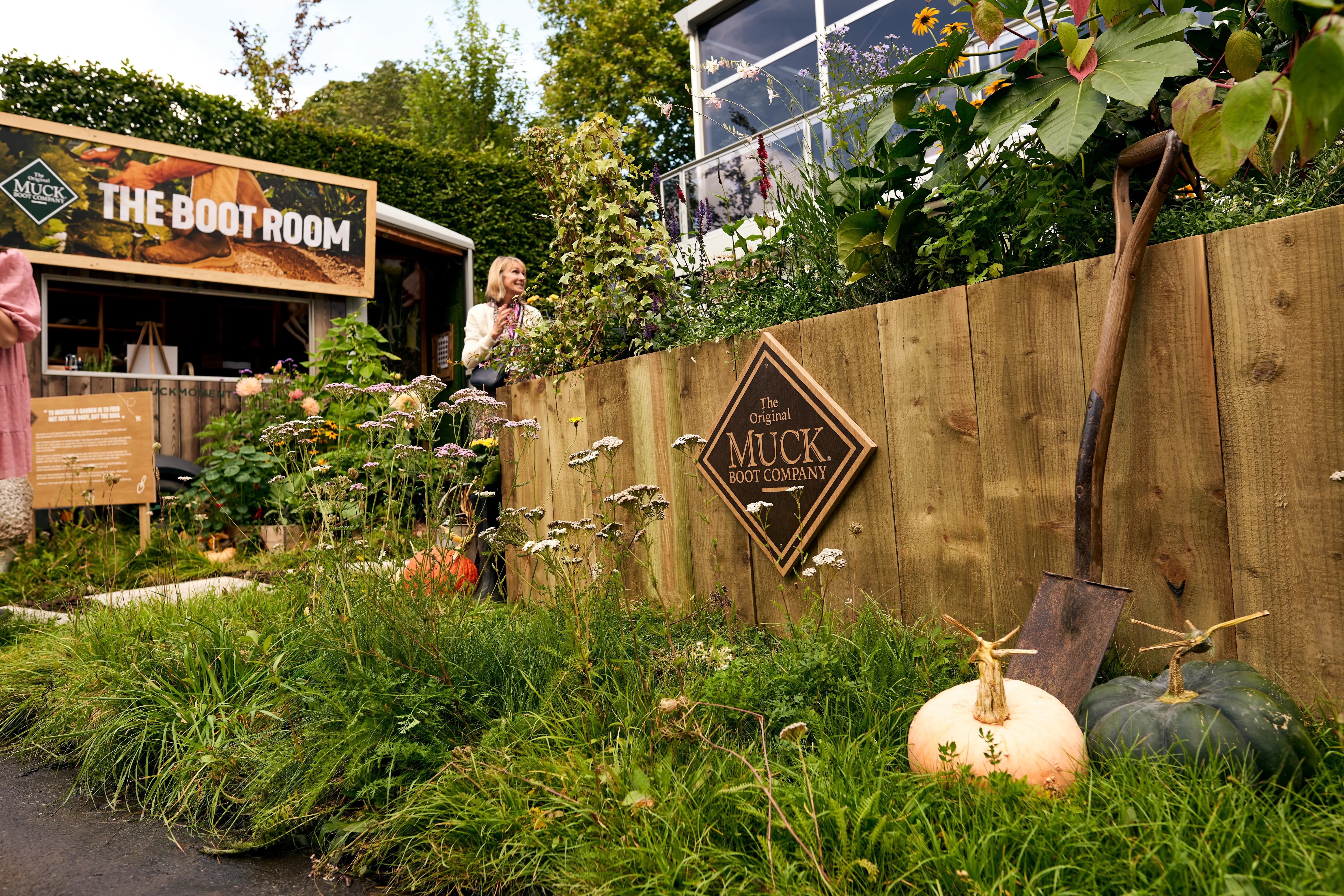 Garden scene featuring a wooden fence, vibrant plants, and a large pumpkin on the ground. A house is in the background.
