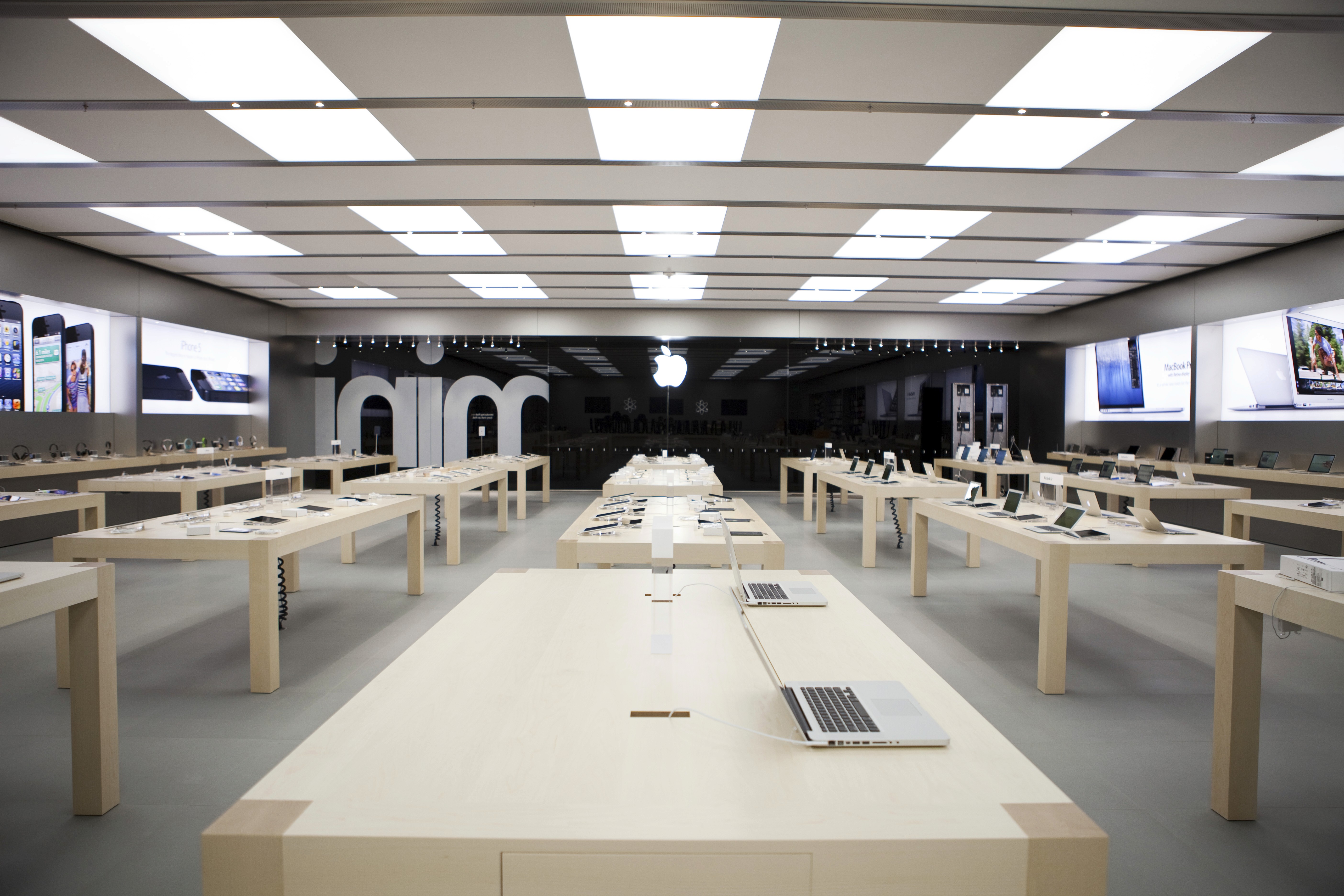 Wide view of an Apple Store interior with long tables and product displays under a bright, geometric ceiling.