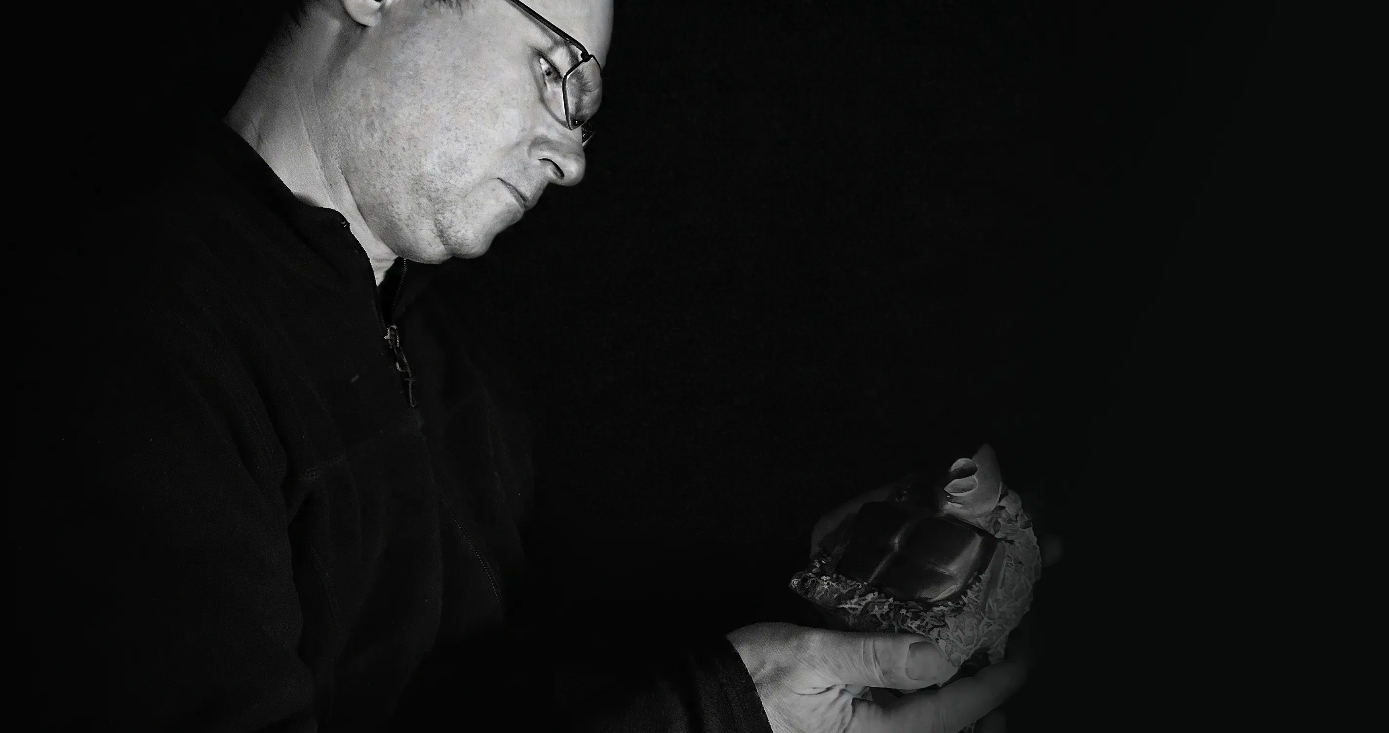 Schwarz-Weiß-Porträt eines Bildhauers mit Brille, der konzentriert eine kleine, handgemißelte Steinskulptur in seinen Händen hält