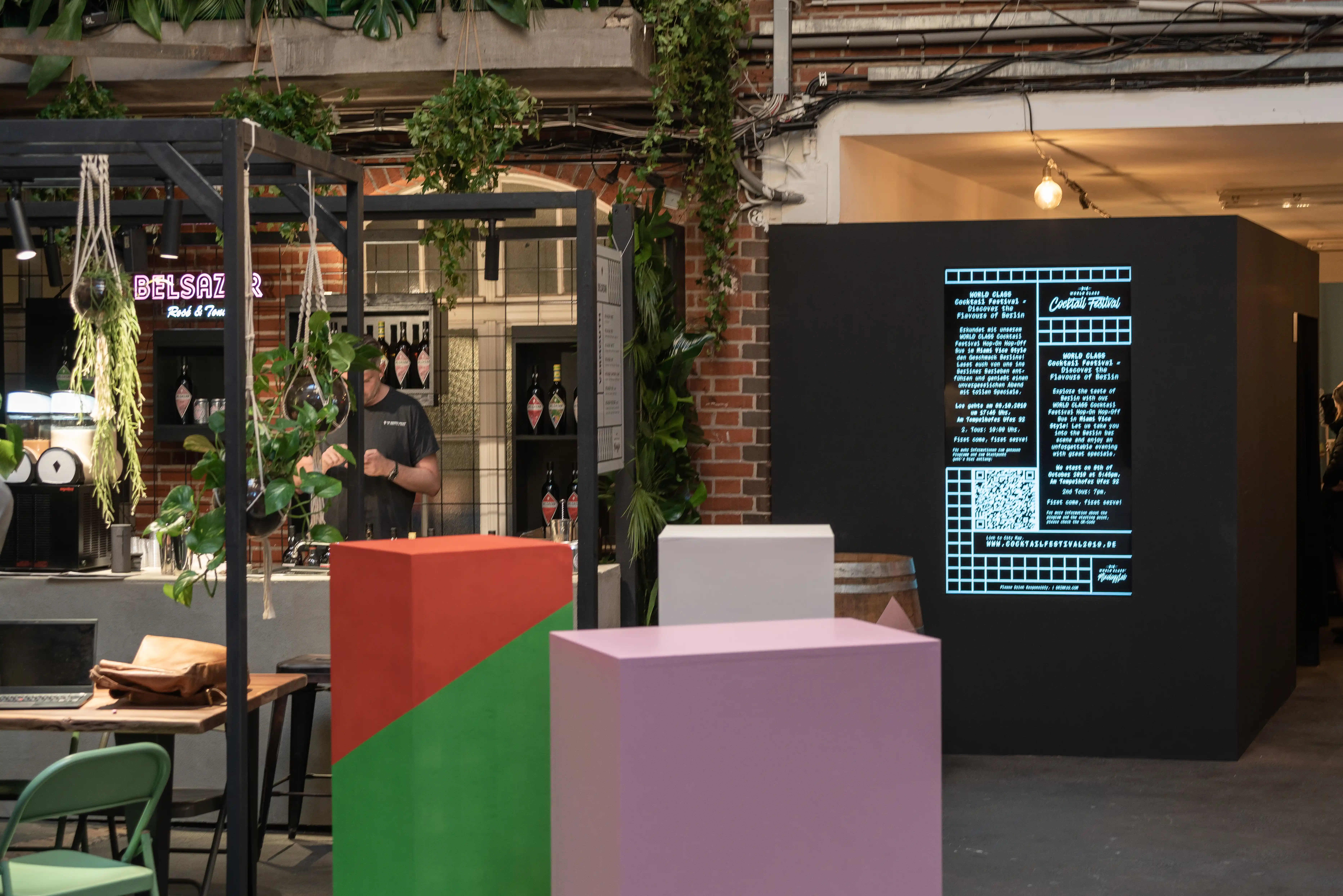 Cocktail area at DIAGEO BCB19 with colorful geometric pedestals, a black wall with blue program text and QR code and the Belsazar bar behind.