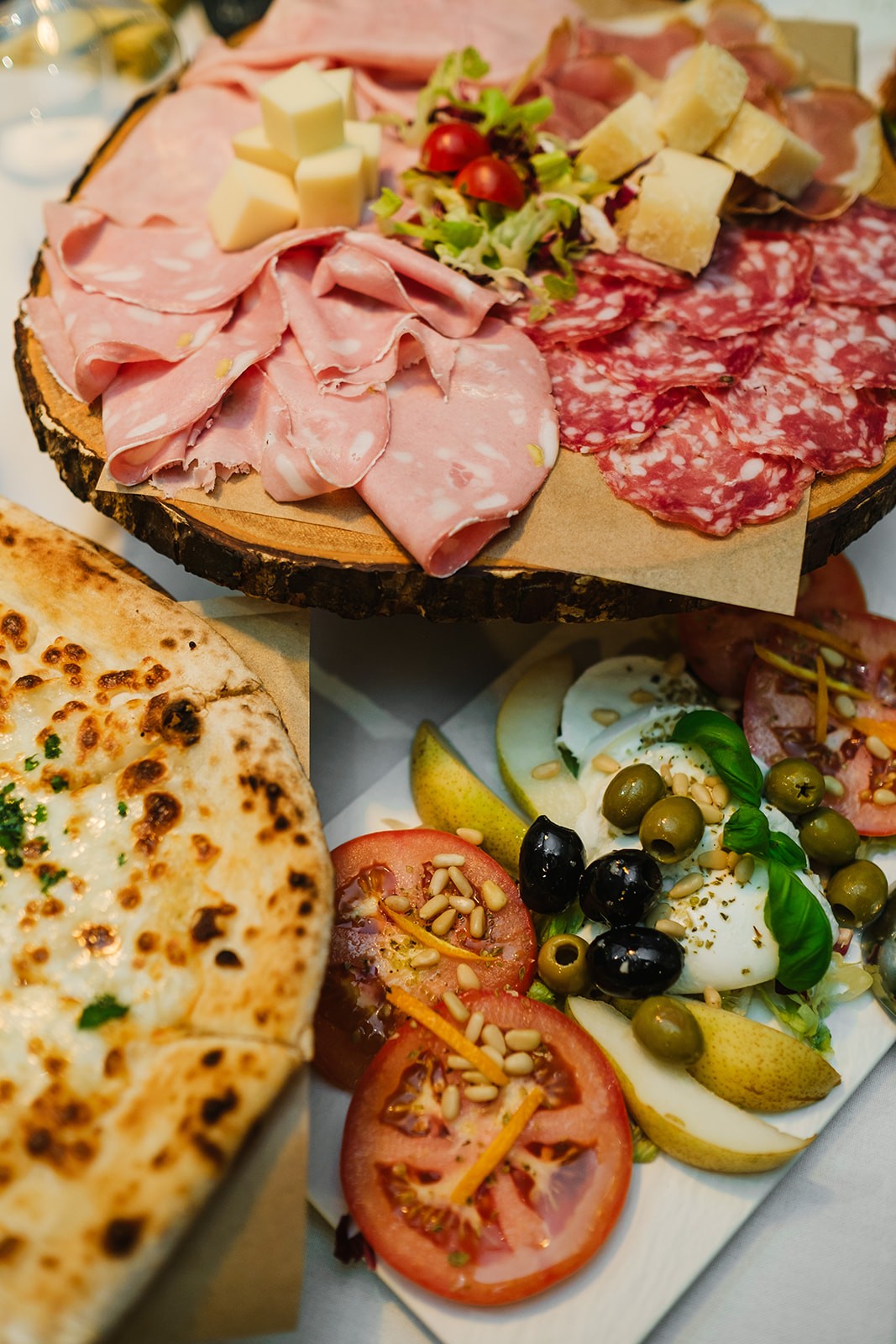 A close-up spread of Italian dishes including a wooden board piled with sliced mortadella, salami, parmesan and salad, a portion of bubbly cheese pizza, and a fresh caprese-style salad with tomatoes, mozzarella, olives, pear slices and pine nuts.