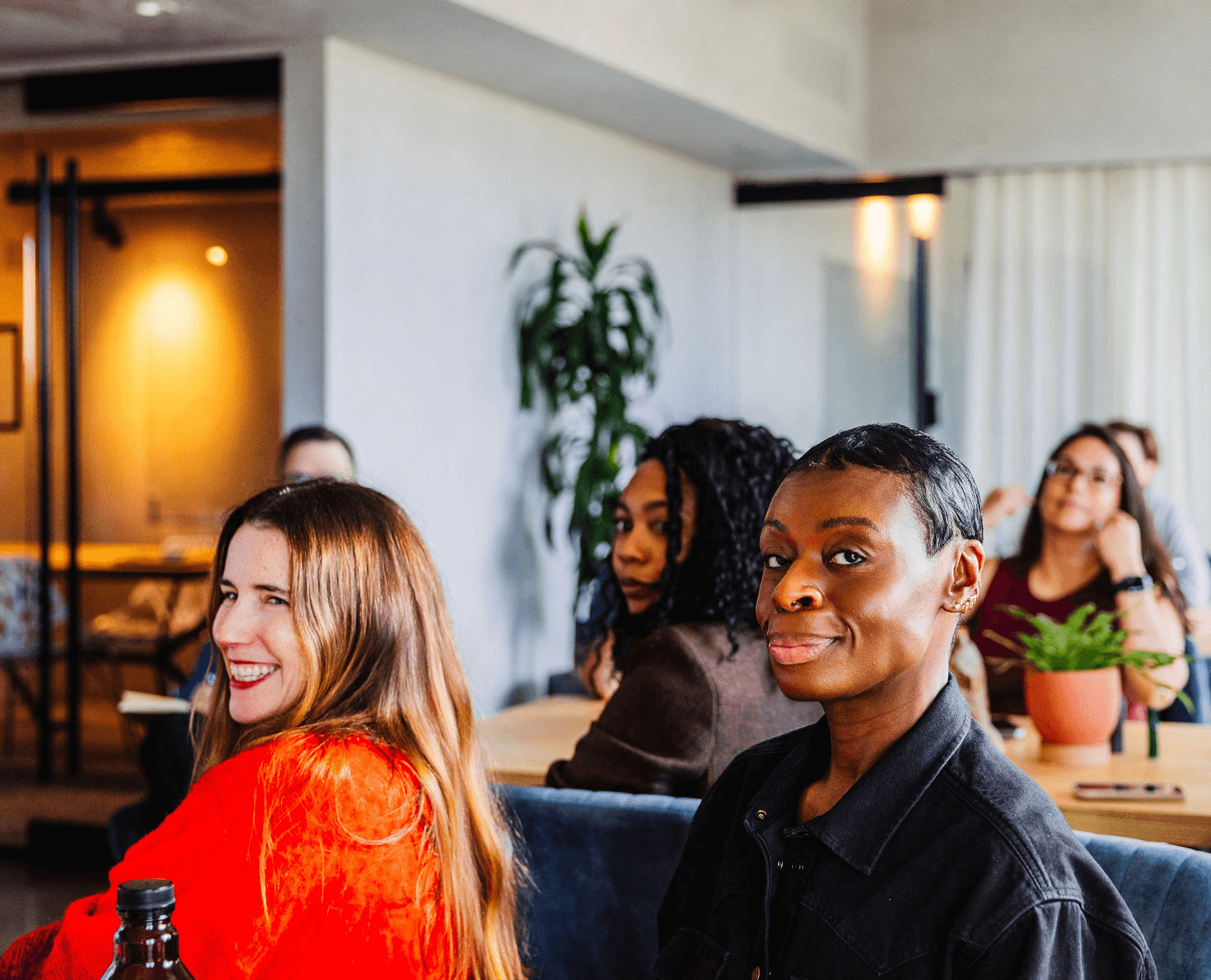 People seated at tables looking toward the camera during a meeting, with notebooks and plants visible in a modern office.