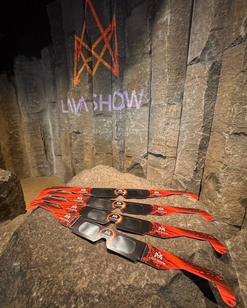 Several pairs of Lava Show-branded solar eclipse glasses with red lava-inspired frames displayed on a large volcanic rock inside Lava Show Reykjavík. The Lava Show logo is projected onto the basalt wall in the background.