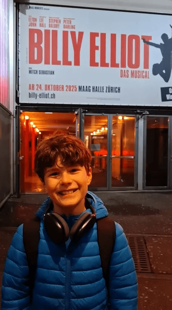 Marius steht strahlend vor dem Plakat des Musicals Billy Elliot in Zürich. Er wurde im Projekt Coaching von Danielle Zuber auf seine Musical-Rolle vorbereitet.
