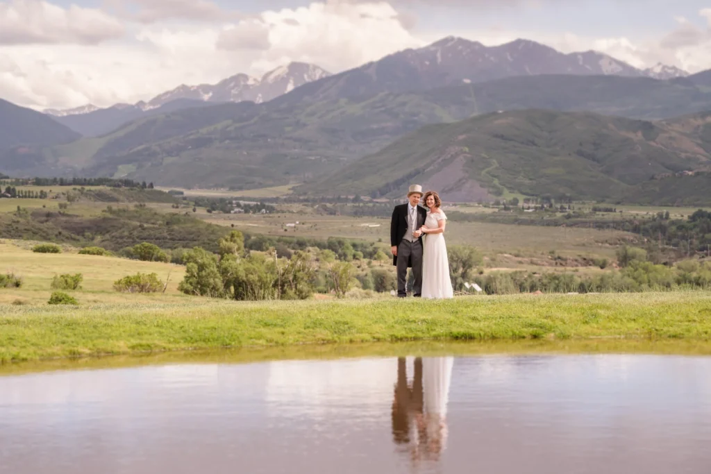 A vertical landscape portrait of the couple standing together on a grassy hill overlooking a vast valley and snow-capped mountains at a Chaparral Ranch Wedding.
