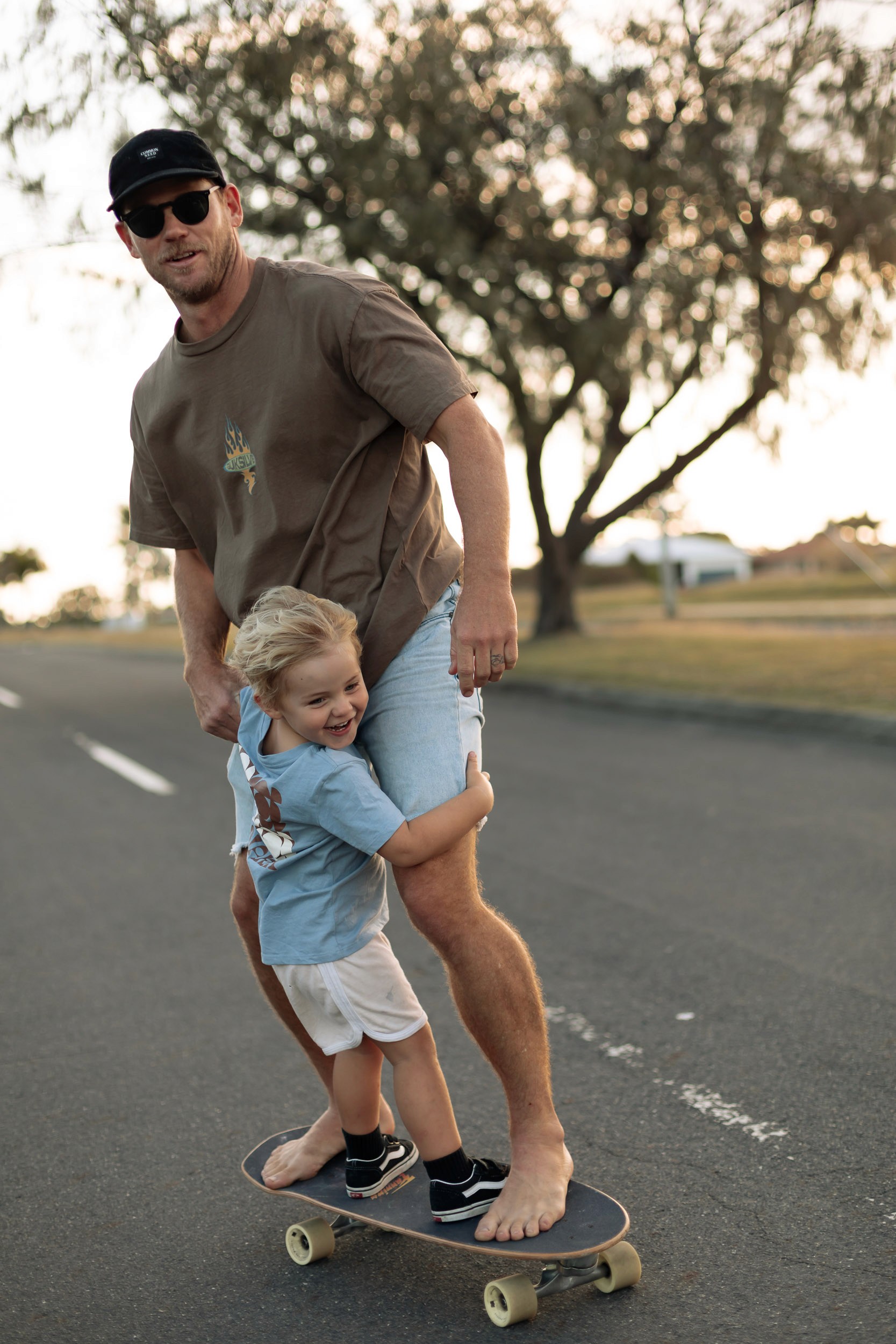 Dad and child riding skateboards together down a sunlit road captured during a candid Mackay family photography session.