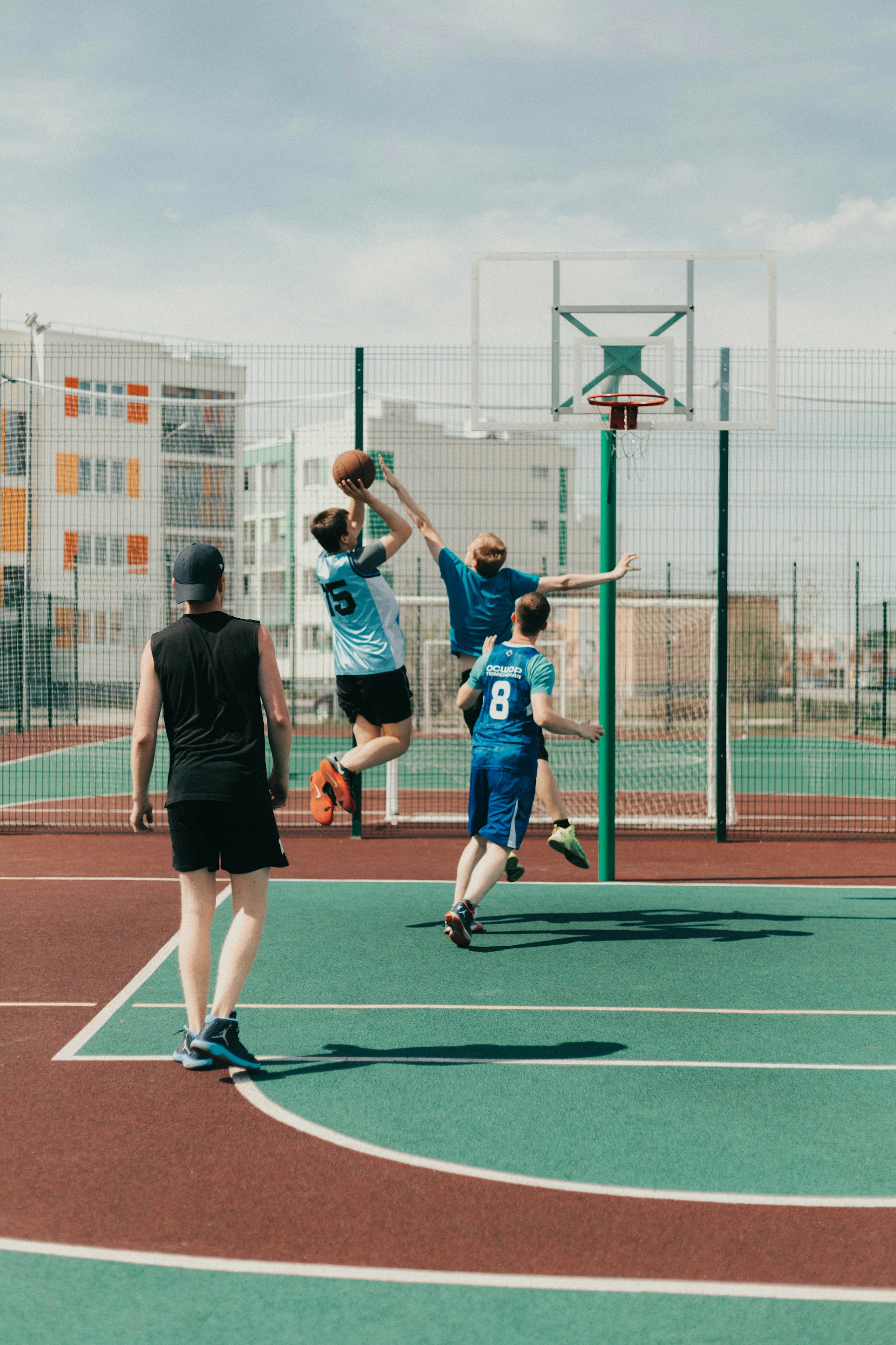 Three young men playing basketball on an outdoor green court with apartment buildings in the background.