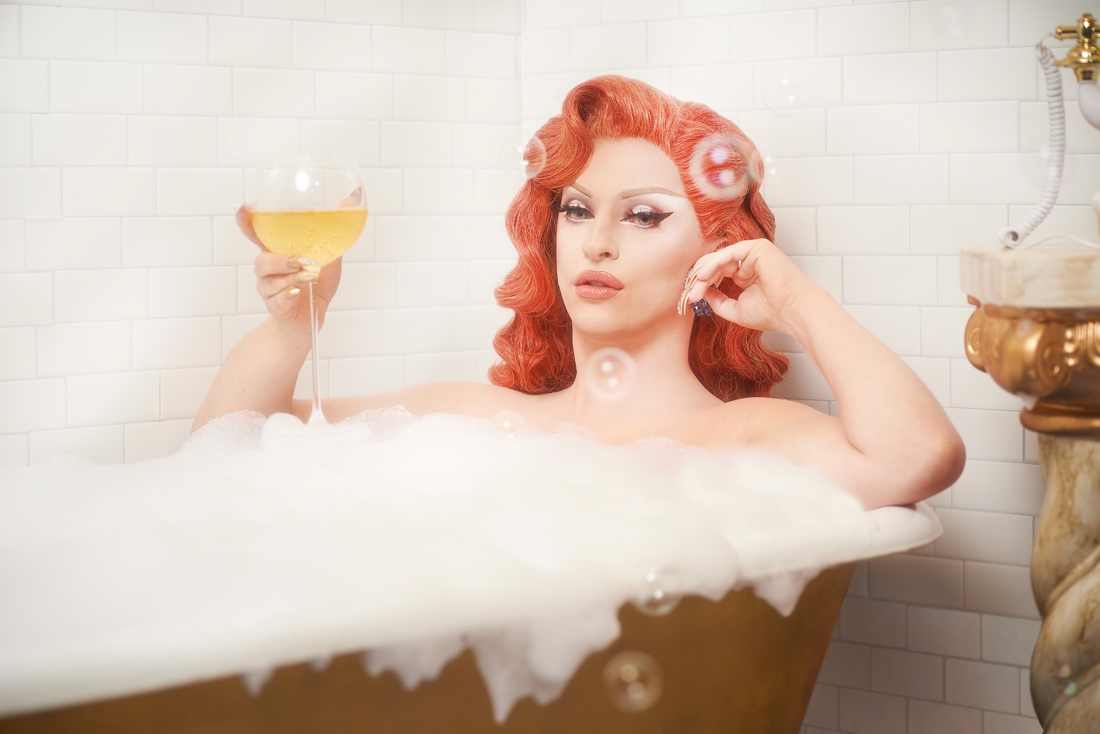 Miz Cracker raising a wine glass while reclining in a foam filled bathtub