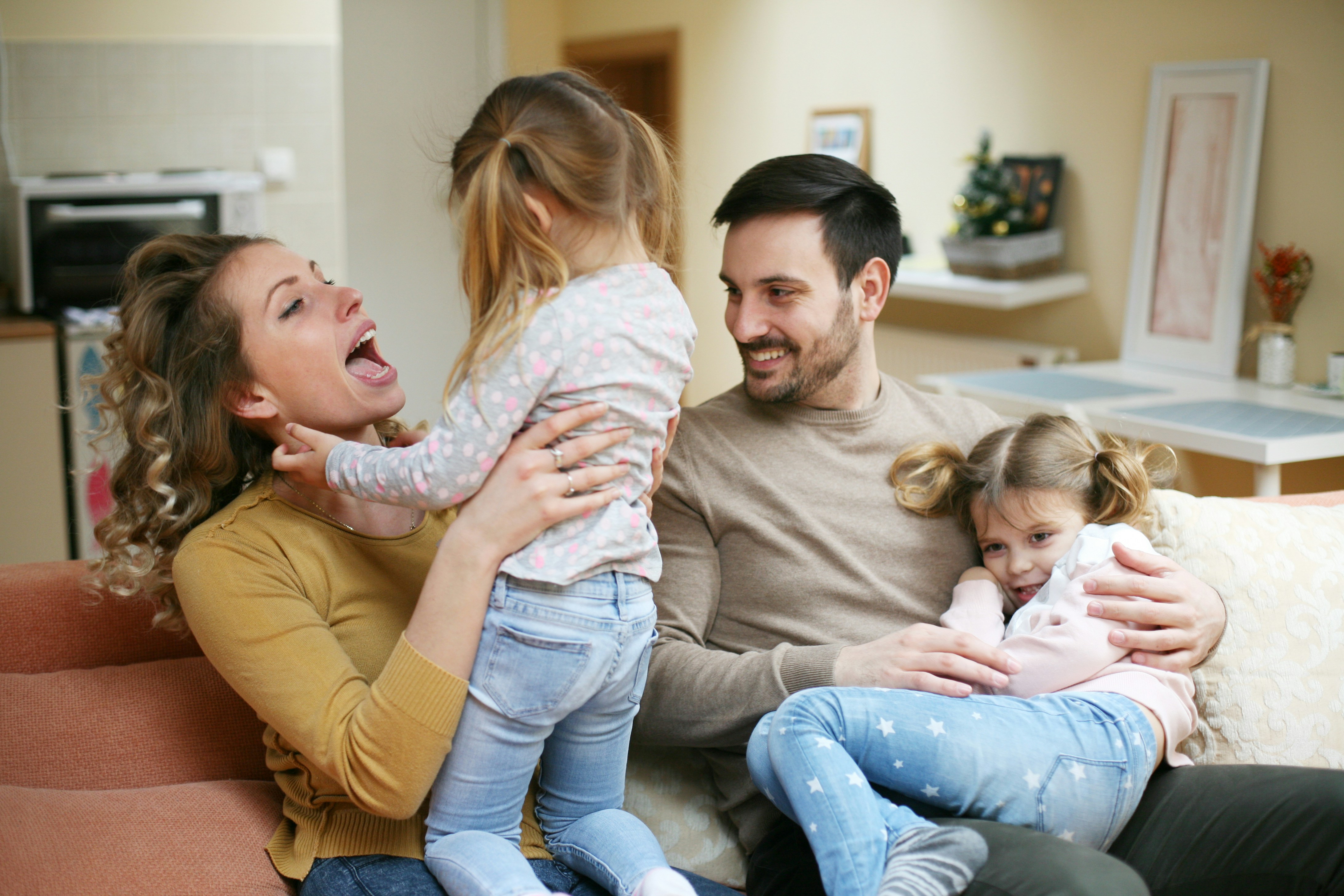 A joyful family of four shares a playful moment in their living room. The mother laughs and interacts with one daughter, while the father holds the other daughter on his lap. The warm, cozy setting highlights the love, happiness, and strong bond within the family.