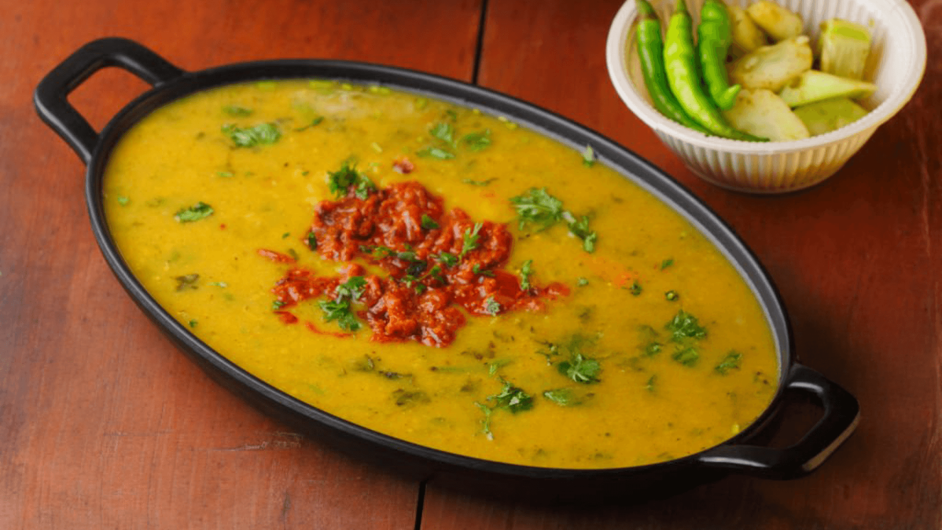 Hot Indian dal garnished with tadka, served in a black dish with fresh green chilies and salad on the side.