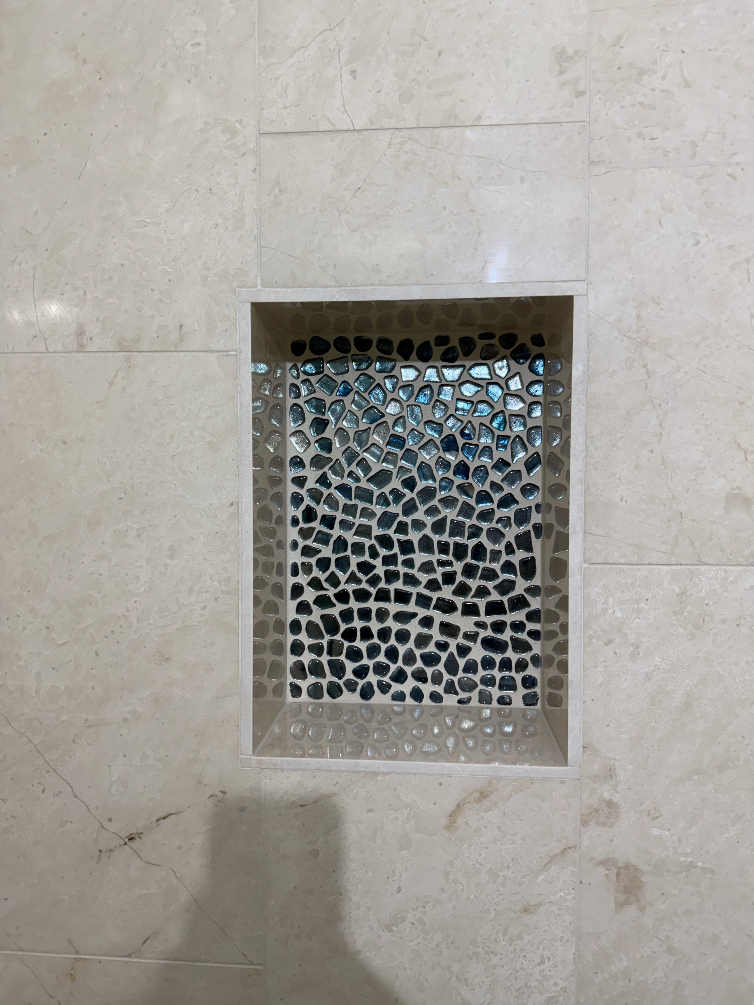 Close-up of a shower niche featuring a vibrant blue and black shattered glass or pebble mosaic tile insert, framed by light beige natural stone wall tiles.
