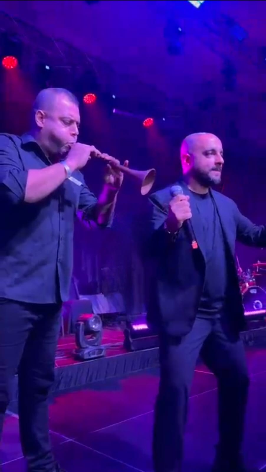 A smiling young woman with curly hair, wearing a purple shirt, stands in a bright indoor setting.