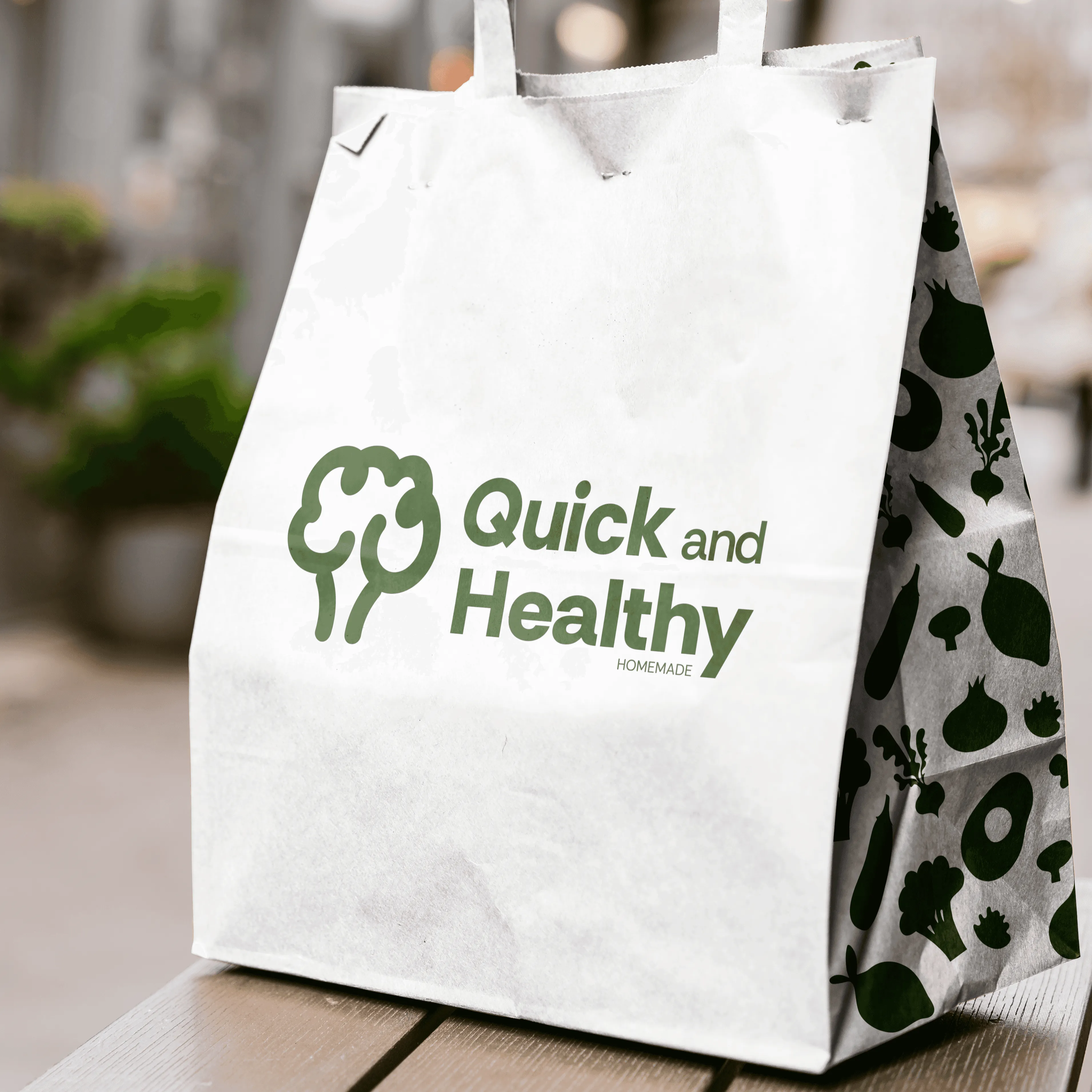 Sac en papier blanc "Quick and Healthy HOMEMADE" avec motifs de légumes verts, posé sur une table en bois à l'extérieur.