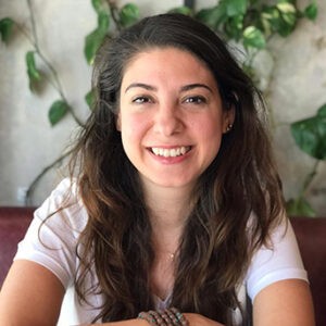 A young person with curly hair smiles at the camera while sitting in front of a laptop against a dark background.