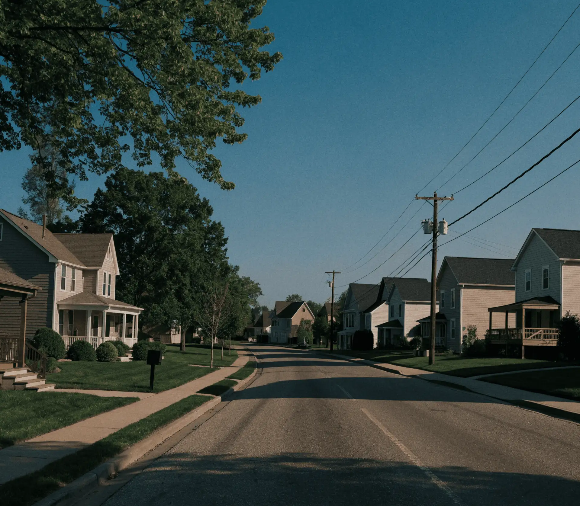 A quiet residential street at dusk lined with two-story clapboard homes, front porches and mature trees